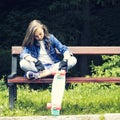Beautiful blonde teen girl in jeans shirt, sitting on bench with backpack and skateboard in park Royalty Free Stock Photo