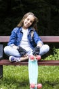 Beautiful blonde teen girl in jeans shirt, sitting on bench with backpack and skateboard in park Royalty Free Stock Photo