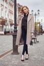 Beautiful blonde girl in autumn coat and handbag looks at the camera adjusting her hair with her free hand