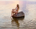 beautiful blonde in a white bikini sits on a stone standing in the water during sunset