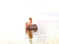 beautiful blonde in a white bikini sits on a stone standing in the water during sunset