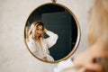 a beautiful blonde in a white shirt looks at her reflection in a round mirror.