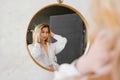 a beautiful blonde in a white shirt looks at her reflection in a round mirror.