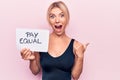 Beautiful blonde woman asking for equality economy holding paper with pay equal message pointing thumb up to the side smiling