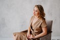 A blonde woman in a beige dress poses on a gray chair in a photo studio.