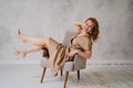 A blonde woman in a beige dress poses on a gray chair in a photo studio.