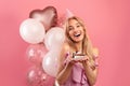 Beautiful blonde woman with birthday hat, balloons and yummy cake looking at camera and smiling on pink background Royalty Free Stock Photo