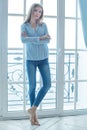Beautiful blonde woman in blue casual denim wear standing in front of window at home Royalty Free Stock Photo