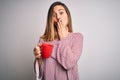 Beautiful blonde woman with blue eyes drinking red mug of coffee over white background cover mouth with hand shocked with shame Royalty Free Stock Photo
