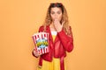 Beautiful blonde woman with blue eyes eating salty popcorn snack over yellow background cover mouth with hand shocked with shame