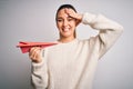 Beautiful blonde woman with blue eyes holding red paper airplane over white background stressed with hand on head, shocked with Royalty Free Stock Photo