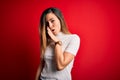 Beautiful blonde woman with blue eyes wearing casual white t-shirt over red background hand on mouth telling secret rumor, Royalty Free Stock Photo