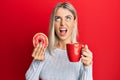 Beautiful blonde woman eating doughnut and drinking coffee angry and mad screaming frustrated and furious, shouting with anger Royalty Free Stock Photo