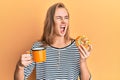 Beautiful blonde woman eating doughnut and drinking coffee angry and mad screaming frustrated and furious, shouting with anger Royalty Free Stock Photo