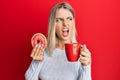 Beautiful blonde woman eating doughnut and drinking coffee angry and mad screaming frustrated and furious, shouting with anger Royalty Free Stock Photo