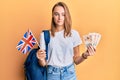 Beautiful blonde woman exchange student holding uk flag and pounds relaxed with serious expression on face