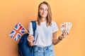 Beautiful blonde woman exchange student holding uk flag and pounds smiling looking to the side and staring away thinking