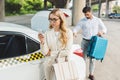 beautiful blonde woman in eyeglasses using smartphone while man putting suitcase in trunk of taxi car