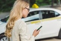 beautiful blonde woman in eyeglasses using smartphone while standing near taxi