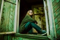 A beautiful blonde woman in a green vintage sweater and jeans is sitting on the windowsill of a wooden abandoned house with broken Royalty Free Stock Photo