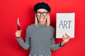 Beautiful blonde woman holding art notebook and brushes sticking tongue out happy with funny expression