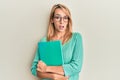 Beautiful blonde woman holding book wearing glasses in shock face, looking skeptical and sarcastic, surprised with open mouth Royalty Free Stock Photo