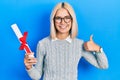 Beautiful blonde woman holding graduate degree diploma smiling happy and positive, thumb up doing excellent and approval sign Royalty Free Stock Photo