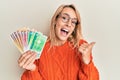Beautiful blonde woman holding norwegian krone banknotes pointing thumb up to the side smiling happy with open mouth