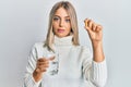 Beautiful blonde woman holding pill and glass of water relaxed with serious expression on face Royalty Free Stock Photo