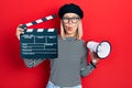 Beautiful blonde woman holding video film clapboard and megaphone making fish face with mouth and squinting eyes, crazy and Royalty Free Stock Photo