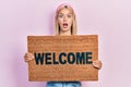 Beautiful blonde woman holding welcome doormat afraid and shocked with surprise and amazed expression, fear and excited face Royalty Free Stock Photo