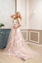 Beautiful blonde woman in pastel floral long puffy dress with makeup and long curly hairstyle standing and posing near dresser at