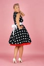 Beautiful blonde woman in a polka-dot dress, coyly turned around over her shoulder and smiling, pin-up style on a pink