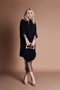 Beautiful blonde woman posing in a black coat with bag on a beige background. Fashion show clothing, woman with perfect figure