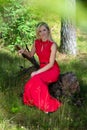 A beautiful, blonde woman in a red long dress sits on a stump in the woods