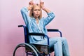 Beautiful blonde woman sitting on wheelchair doing funny gesture with finger over head as bull horns