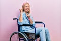 Beautiful blonde woman sitting on wheelchair pointing to the back behind with hand and thumbs up, smiling confident Royalty Free Stock Photo