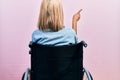 Beautiful blonde woman sitting on wheelchair posing backwards pointing ahead with finger hand Royalty Free Stock Photo