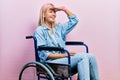 Beautiful blonde woman sitting on wheelchair very happy and smiling looking far away with hand over head Royalty Free Stock Photo