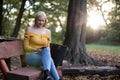 Beautiful blonde woman sitting on a wooden bench outdoors in the park, using smartphone Royalty Free Stock Photo