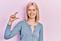 Beautiful blonde woman standing over pink background smiling and confident gesturing with hand doing small size sign with fingers Royalty Free Stock Photo