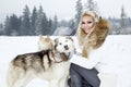 Beautiful blonde woman, standing on snow and holding Husky dogs view of mountains and snow.