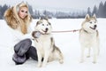 Beautiful blonde woman, standing on snow and holding Husky dogs view of mountains and snow.