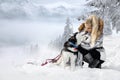 Beautiful blonde woman, standing on snow and holding Husky dogs view of mountains and snow.