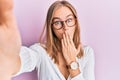 Beautiful blonde woman taking a selfie photo wearing glasses covering mouth with hand, shocked and afraid for mistake