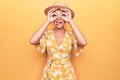 Beautiful blonde woman on vacation wearing summer hat and dress over yellow background doing ok gesture like binoculars sticking Royalty Free Stock Photo