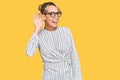 Beautiful blonde woman wearing business shirt and glasses smiling with hand over ear listening an hearing to rumor or gossip Royalty Free Stock Photo