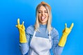 Beautiful blonde woman wearing cleaner apron and gloves shouting with crazy expression doing rock symbol with hands up