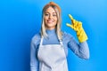 Beautiful blonde woman wearing cleaner apron and gloves smiling and confident gesturing with hand doing small size sign with Royalty Free Stock Photo