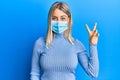 Beautiful blonde woman wearing covid-19 medical mask smiling with happy face winking at the camera doing victory sign